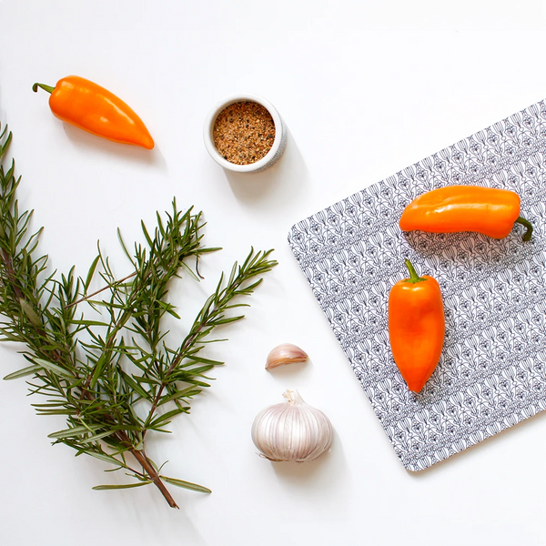 Leikkuulauta ja pala-minikuppi jossa maustetta. Leikkuulaudassa Sirkus-musta kuosi. Laudan päällä on kaksi oranssia suippoparikaa ja pöydällä yksi sekä rosmaariinin oksa ja valkosipulin puolikas, josta irroitettu yksi kynsi. Tausta on valkoinen. Kuva on yläperspektiivistä.